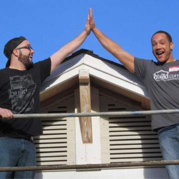 Two smiling men standing on a scaffolding in front of the roof of a home, their hands together in a high five.