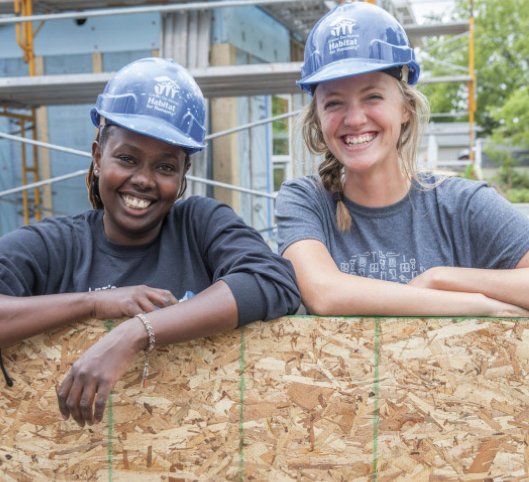 Two women volunteers, smiling with hard hats on, standing in front of a scaffolding, and leaning on a wooden wall.