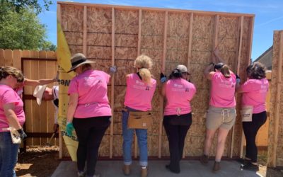 A group of women with their backs to us, holding up a wall that is being built.