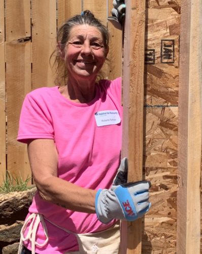 A volunteer posing and smiling with work gloves on, next to a home construction in progress.