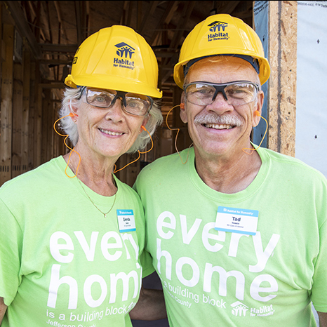 Two volunteers, a man and a woman, with hard hats on, smiling in front of a building.