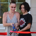 Abbey and one other person next to her, standing in front of a home, cutting a red ribbon.